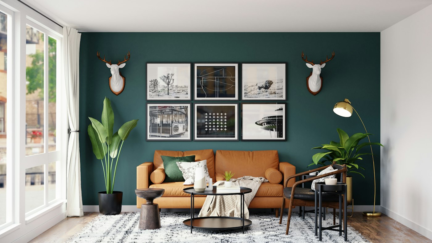 Tan leather sofa on a dark green wall, six framed pictures, two white deer heads, and a brass lamp.
